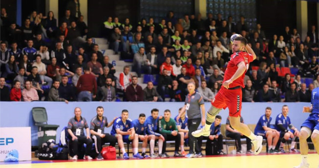 Në foto shihet një lojtar me top në dorë i cili është hedhur lartë për të gjuajtur në portën e kundërshtarit.