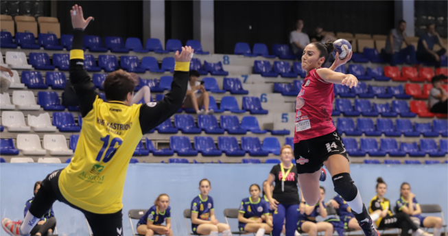 Një lojtare me top në dorë është hedhur lartë për të shënuar gol, ndërsa portierja kundërshtare tenton ta ndalojë me duart lartë.