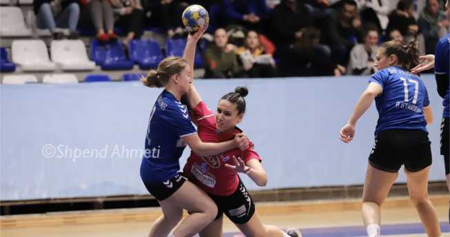 Lojtarja ka topin në dorë dhe po mundohet të shënojë gol, kurse kundërshtarja e ka kapur për fanellë duke tentuar ta ndalojë.