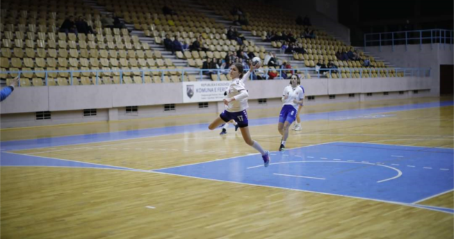 Në foto shihet një lojtare e cila në dorë ka topin dhe është hedhur lartë për të shënuar gol, kurse mbrapa është një bashkëlojtare e saj duke vrapuar.