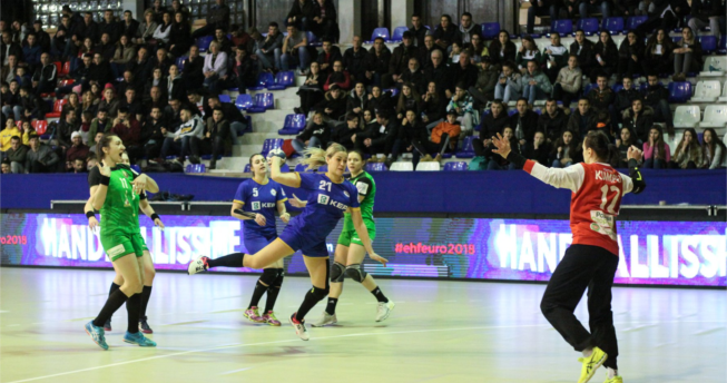 Në foto shihet një lojtare e Kombëtares sonë me top në dorë duke u munduar të shënojë gol, mbrapa saj është një bashkëlojtare, dhe tri lojtare te ekipit kundërshtar, kurse përballë ka portieren e ekipit kundërshtar duke tentuar ta bllokoje gjuajtjen e saj.