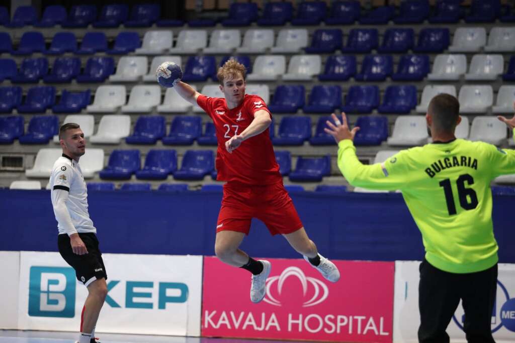 Great Britain player wearing red shirt in mid air with ball in hand shooting at goal